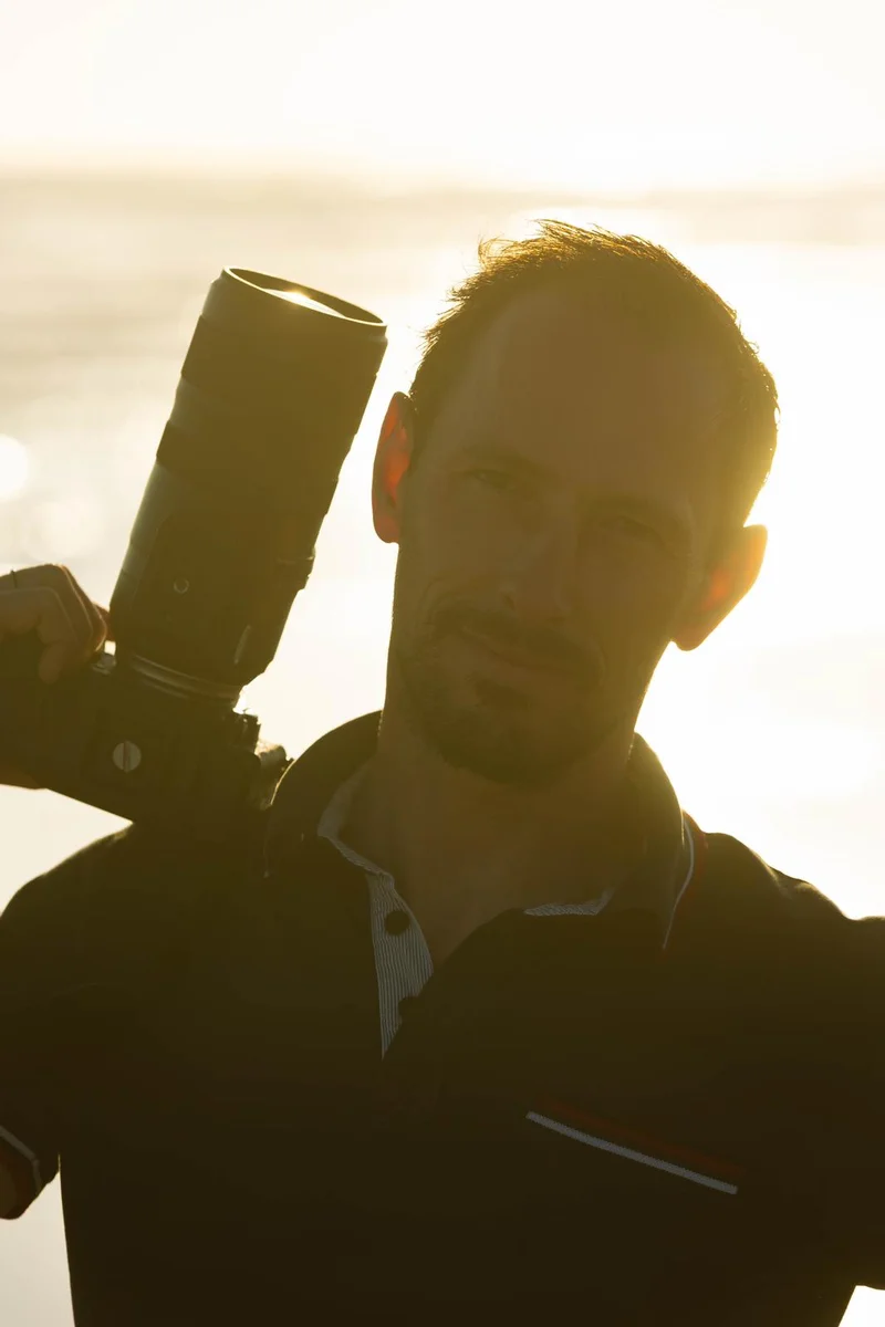 Portrait d'Aurélien Lambert tenant son appareil photo à l'épaule, éclairé en contre-jour par la lumière dorée du soleil
